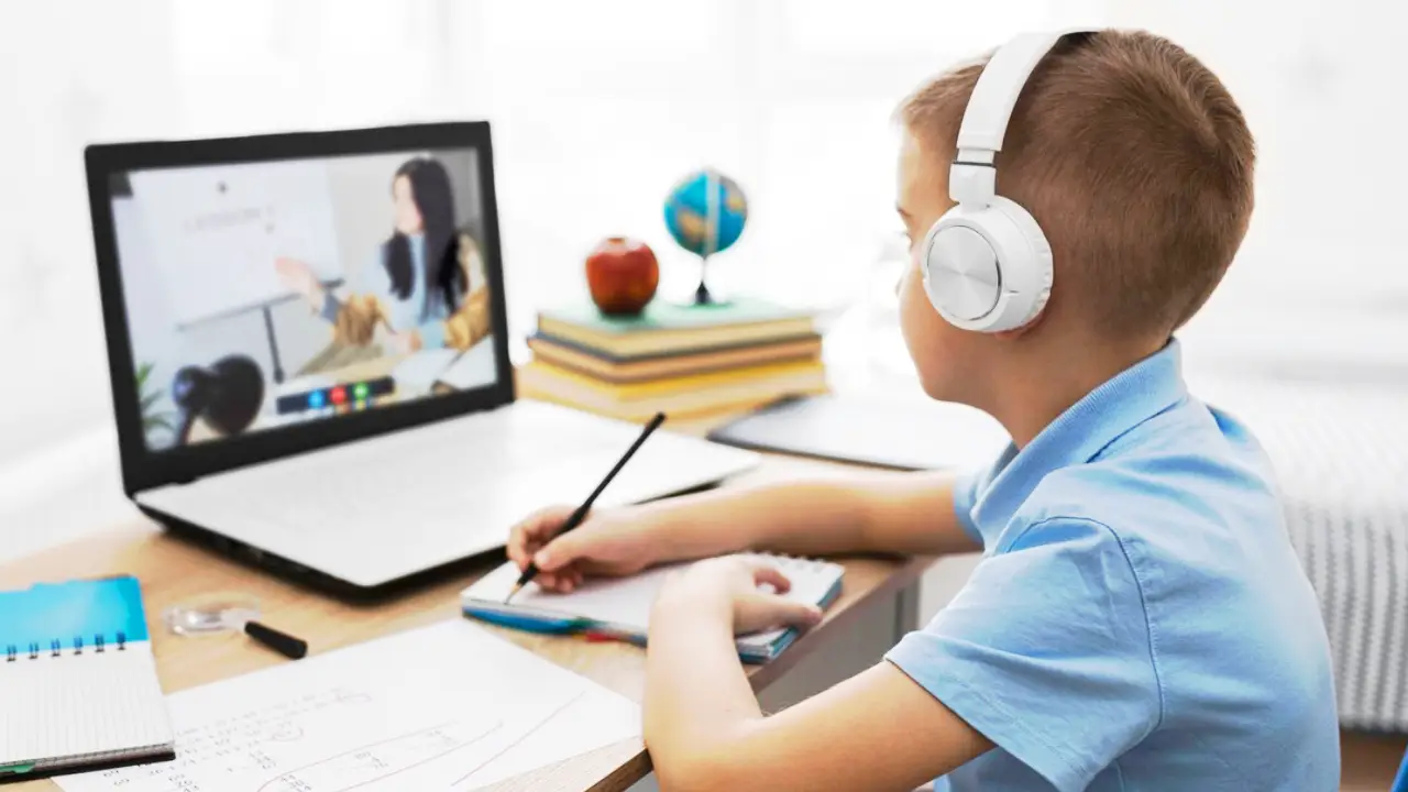少年がオンラインレッスンを受けています。机に座り、コンピューターに向かってヘッドフォンを着け、メモを取っています。 A young boy is having an online lesson. He is sitting at a desk, facing a computer, wearing headphones, and taking notes.